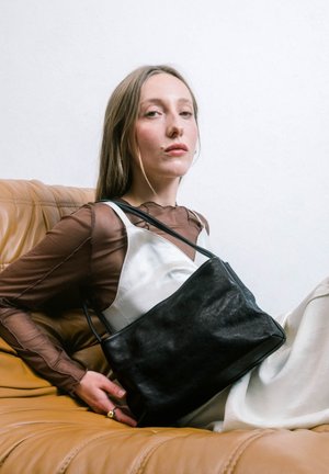 Mujer con cabello largo y rubio que lleva una blusa marrón translúcida y un vestido blanco, sentada en un sofá de cuero marrón claro, sosteniendo un gran bolso de cuero negro.