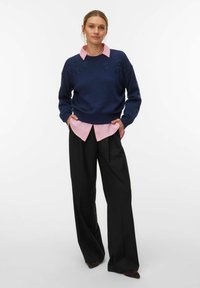 Femme debout les mains dans les poches, portant un pull bleu marine à motifs floraux sur une chemise rose et un pantalon large noir avec des chaussures à bouts pointus.