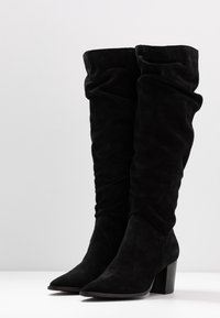Bottes noires en suede au-dessus du genou avec un design froncé, bout pointu et talon épais, présentant une texture douce et une finition lisse.