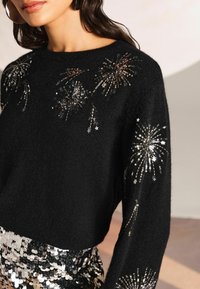 Czarny krótki sweter z naszytym wzorem fajerwerków z cekinów na rękawach, z metalicznymi niteczkami i miękką teksturą. W zestawie z cekinową spódnicą.
