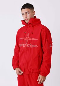 Jonge man in een rode hooded jacket en broek met het witte PXLP-logo, staand tegen een effen lichte achtergrond.