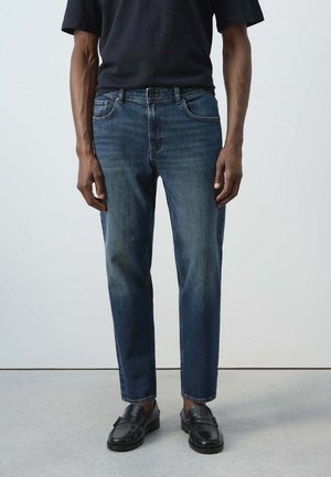 Mann mit dunkelblauer Jeans, schwarzen Schuhen und schwarzem T-Shirt steht vor einem einfarbigen weißen Hintergrund.