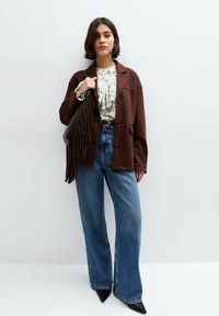 Giacca di camoscio marrone, blusa floreale, jeans a vita alta svasati blu, tacchi a punta neri, porta una borsa a tracolla nera con frange.