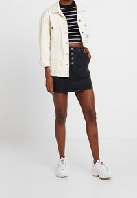 Jupe en jean noir mini avec boutons à l'avant, associée à un haut court noir et blanc à rayures et une veste en jean crème. Baskets blanches.