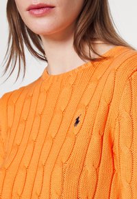 Nainen, jolla on päällään oranssi palmikkoneulepaita, jossa on pieni laivastonsininen polo-pelaajan logo rinnassa.