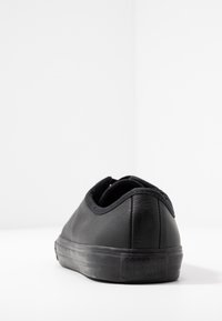 Chaussure Converse noire en cuir, basse, vue de dos sur une surface blanche avec un arrière-plan minimaliste.