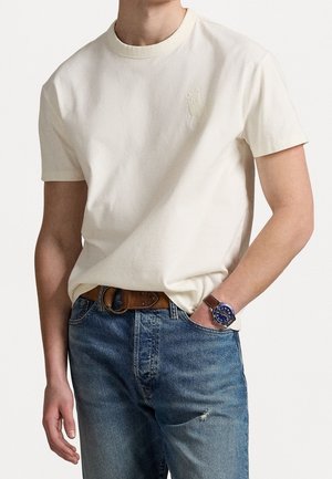 Cremefarbenges T-Shirt mit kurzen Ärmeln und rundem Ausschnitt sowie gesticktem Logo. Kombiniert mit blauen Jeans aus Denim und einem braunen Ledergürtel.