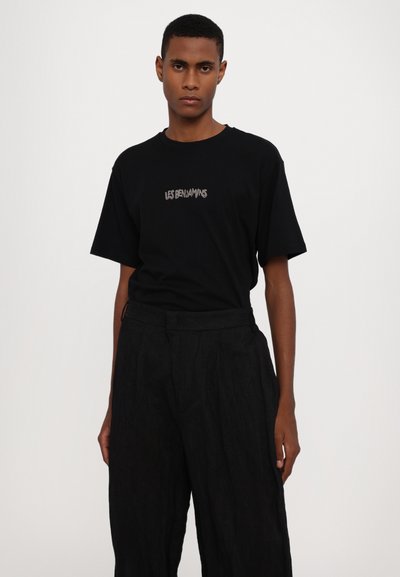 Camiseta negra de manga corta con un logo gráfico "LES BENJAMINS" en el pecho, combinada con pantalones negros holgados. Diseño simple y minimalista.