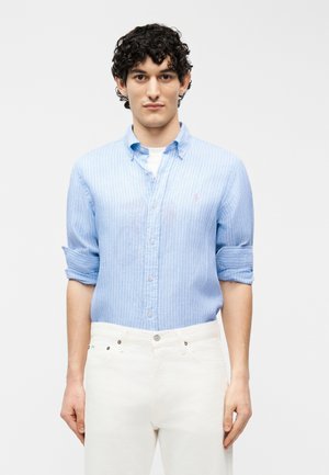 Młody mężczyzna z krótkimi kręconymi włosami ubrany w jasnoniebieską, pasiastą koszulę z podwiniętymi rękawami i białe spodnie, stojący na białym tle.