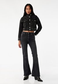 Sort cropped denimjakke med slidte detaljer, knaplukning foran og to brystlommer, parret med sorte flared jeans og støvler med hæl.