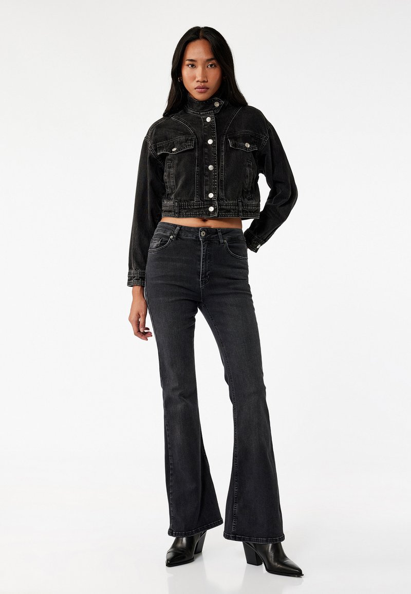 Sort cropped denimjakke med slidte detaljer, knaplukning foran og to brystlommer, parret med sorte flared jeans og støvler med hæl.