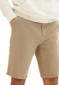 Beige Shorts aus strukturiertem Stoff. Verfügt über schräge Seitentaschen, einen Reißverschluss vorne und umsäumte Saumabschlüsse. Getragen mit einem hellen Kapuzenpullover.