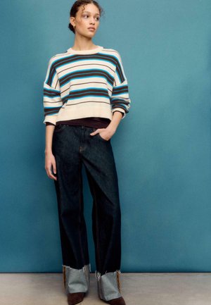 Gestreifter Strickpullover in Creme, Braun und Blau. Dunkelblaue Denim-Hose mit weitem Bein und ausgefranstem Saum. Braune Stiefeletten mit grauen Akzenten.