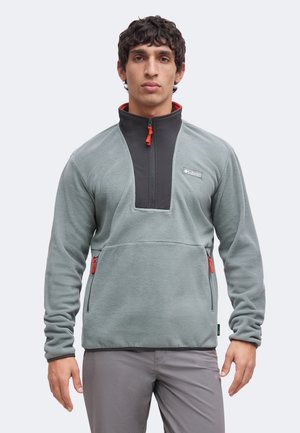 Mand iført en grå fleece-pullover med sort høj krave og orange lynlåstræk, stående mod en ensartet lys baggrund.