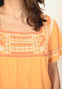 Oranžs tunika ar sarežģītu baltu un rozā izšuvumu ap kvadrātveida dekoltē un piedurknēm, brīvā, plūstošā piegriezumā un mīkstā tekstūrā.