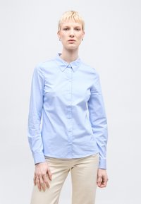 Άτομο με κοντά ξανθά μαλλιά, φορούν ένα ανοιχτό γαλάζιο πουκάμισο με κουμπιά και μπεζ παντελόνι, στέκεται μπροστά σε απλό λευκό φόντο.