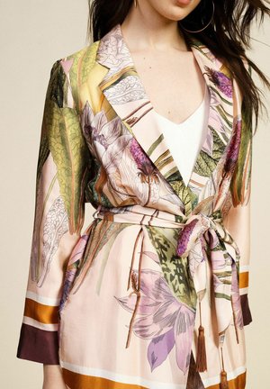 Donna che indossa un blazer di seta rosa floreale con foglie verdi, fiori viola, cintura annodata in vita e una maglia bianca sotto.