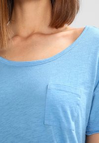 Světle modré tričko s krátkým rukávem vyrobené z jemného materiálu, s kulatým výstřihem a malou přišitou kapsičkou na levé hrudi.