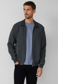 Graue, strukturierte Reißverschlussjacke mit Kragen, versehen mit gerippten Bündchen und Saum, über einem hellblauen T-Shirt getragen. Dunkle Hose vervollständigt das Outfit.