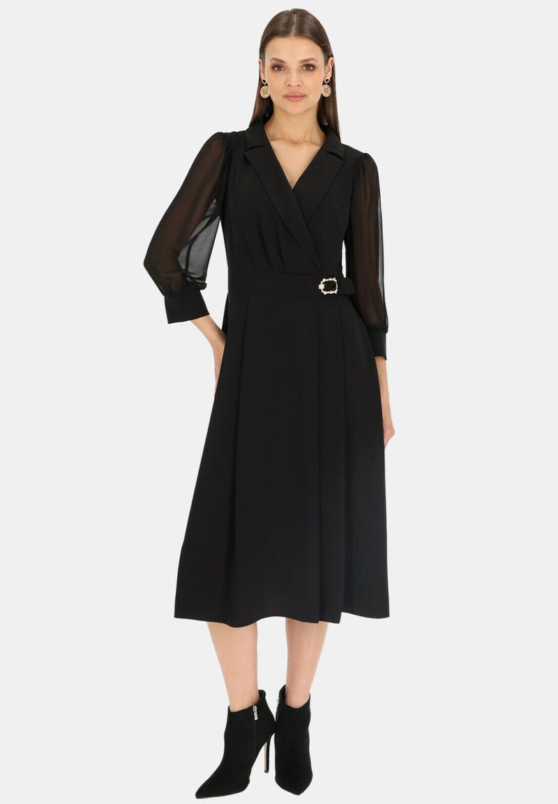 Czarna sukienka w stylu wrap z przezroczystymi, długimi rękawami, przewiązana w talii złotą klamrą. Sięga poniżej kolan. Noszona z czarnymi botkami za kostkę.