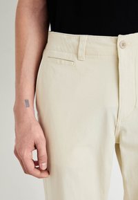 Pantaloni din bumbac bej cu o textură fină, având închidere cu nasture, buzunare laterale și un design slim fit; mână odihnindu-se pe țesătură.