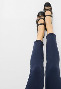 Schwarze Ballettschuhe aus Mesh mit quadratischen Zehen und doppelten Riemen, kombiniert mit dunkelblauen, taillierten Hosen. Glatte Textur und schlankes Design.