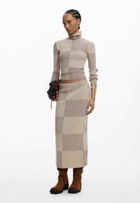 Kobieta w beżowo-brązowym prążkowanym swetrze z golfem i dopasowanej długiej spódnicy, trzymająca małą ciemną torebkę i nosząca brązowe kozaki.