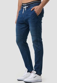 Pantalones jogger de mezclilla con un ajuste slim, color azul oscuro, cinturilla elástica con cordón, bolsillos laterales y detalles de costura. Combinados con zapatillas blancas.