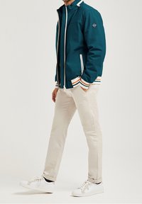 Teal jacka med vit dragkedja och randiga ärmslut i orange och grönt. Matchad med ljusbeiga byxor och vita sneakers.