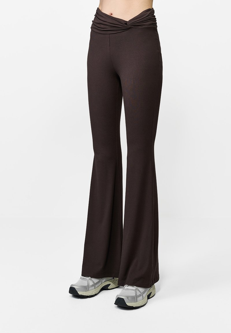 Brązowe, rozszerzane spodnie z skręconym detalem w pasie, wykonane z miękkiego, elastycznego materiału. W połączeniu z szarymi butami sportowymi.