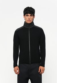 Μαύρο ζιβάγκο πουλόβερ με φερμουάρ, ψηλό γιακά, υφασμάτινους ώμους με υφή, εφαρμοστά μανίκια και ριμπ τζιν στο κάτω μέρος, κατασκευασμένο από απαλό μείγμα υφασμάτων.