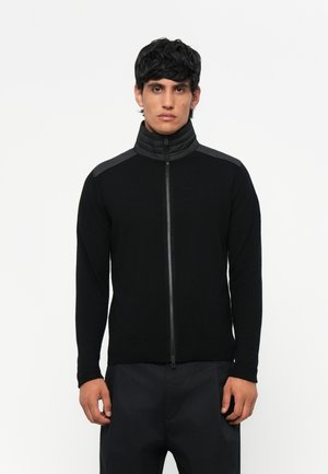 Sudadera negra con cremallera y cuello alto, con detalles texturizados en los hombros, mangas ajustadas y dobladillo acanalado, confeccionada en una mezcla de telas suaves.