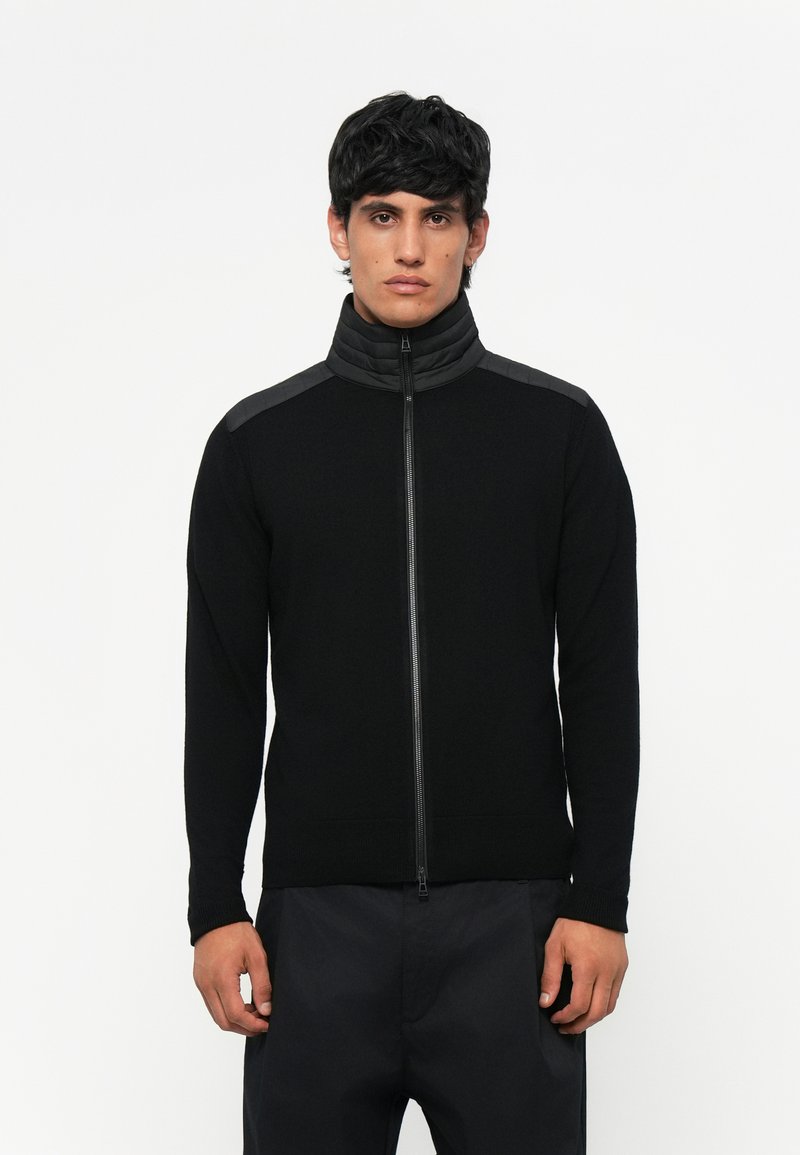 Μαύρο ζιβάγκο πουλόβερ με φερμουάρ, ψηλό γιακά, υφασμάτινους ώμους με υφή, εφαρμοστά μανίκια και ριμπ τζιν στο κάτω μέρος, κατασκευασμένο από απαλό μείγμα υφασμάτων.