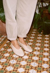Scarpe slip-on in pelle beige con punte appuntite, indossate con pantaloni chiari arrotolati, su un pavimento piastrellato a motivi.