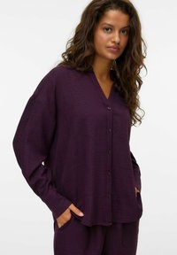 Chemise violette à boutons avec un tissu texturé, design en v, épaules tombantes et poches latérales. La chemise est à coupe ample et à manches longues.