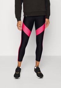 Person trägt schwarze und pinke Sportleggings, schwarze Turnschuhe und ein dunkles Langarmoberteil. Sie steht vor einem hellen Boden und einer hellen Wand.