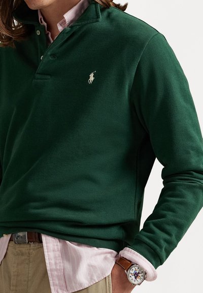 Polo en coton vert à manches longues avec un col et trois boutons, arborant un petit logo blanc. Porté par-dessus une chemise rayée rose clair.