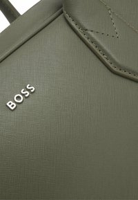 Nahaufnahme einer strukturierten olivgrünen Tasche mit genähtem Detail und silbernen Buchstaben, die BOSS auf der Oberfläche buchstabieren.