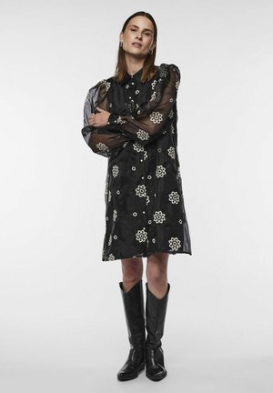 Schwarzes Kleid mit floralem Stickmuster, durchsichtigen langen Ärmeln und einer Knopfleiste. Kombiniert mit kniehohen schwarzen Stiefeln.
