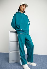 En teal hettegenser med frontlomme, hvite striper på ermene, matchende teal bukser med hvite sidestiper, og hvite sneakers.