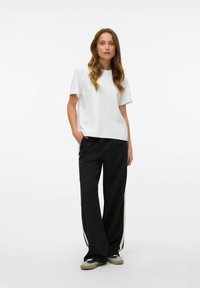 Witte t-shirt met korte mouwen, gecombineerd met zwarte, wijde broek met crèmekleurige zijstrepen en grijze sneakers. Soepele stof en een relaxte pasvorm.
