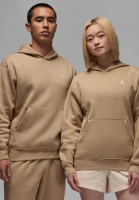 Felpe con cappuccio color sabbia, dotate di tasca a marsupio e logo ricamato, realizzate in un tessuto liscio. I modelli indossano pantaloni coordinati, uno dei quali è un paio di pantaloni corti.