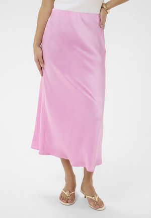 Femme portant une jupe midi en satin rose, un haut blanc, des sandales beige à talons bas avec des détails dorés, et un bracelet doré au poignet.