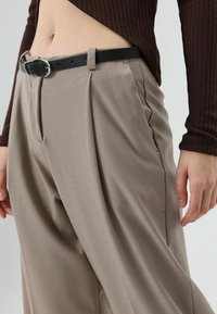 Pantalon plissé beige à taille haute, confectionné dans un tissu lisse, associé à une ceinture en cuir noire avec une boucle argentée.