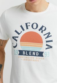 Baltas medvilnės marškinėliai su grafiniu dizainu, kuriame pavaizduota didelė oranžinė saulė, mėlynos juostos ir tekstas "California Blend 1993."