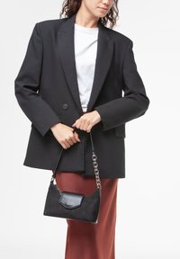 Borsa a tracolla in suede nero con manico a catena argentata e chiusura a pattina, tenuta da una persona che indossa un blazer nero e una gonna marrone.