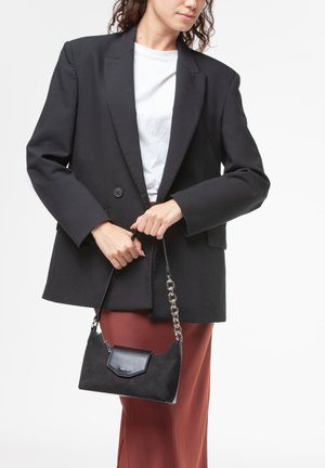 Borsa a tracolla in suede nero con manico a catena argentata e chiusura a pattina, tenuta da una persona che indossa un blazer nero e una gonna marrone.