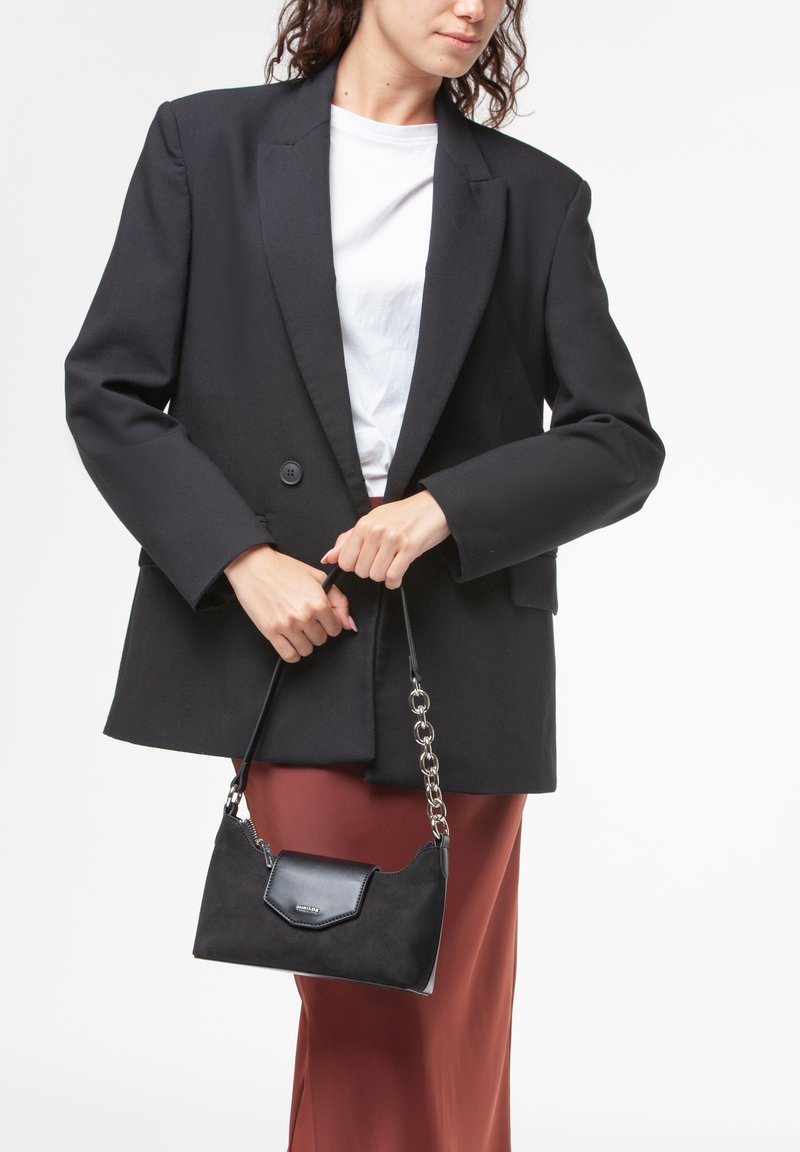 Borsa a tracolla in suede nero con manico a catena argentata e chiusura a pattina, tenuta da una persona che indossa un blazer nero e una gonna marrone.