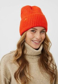 Beanie arancione lavorato a maglia con texture a coste, caratterizzato da un orlo piegato. Indossato con un maglione beige a collo alto, che mette in mostra un look casual.