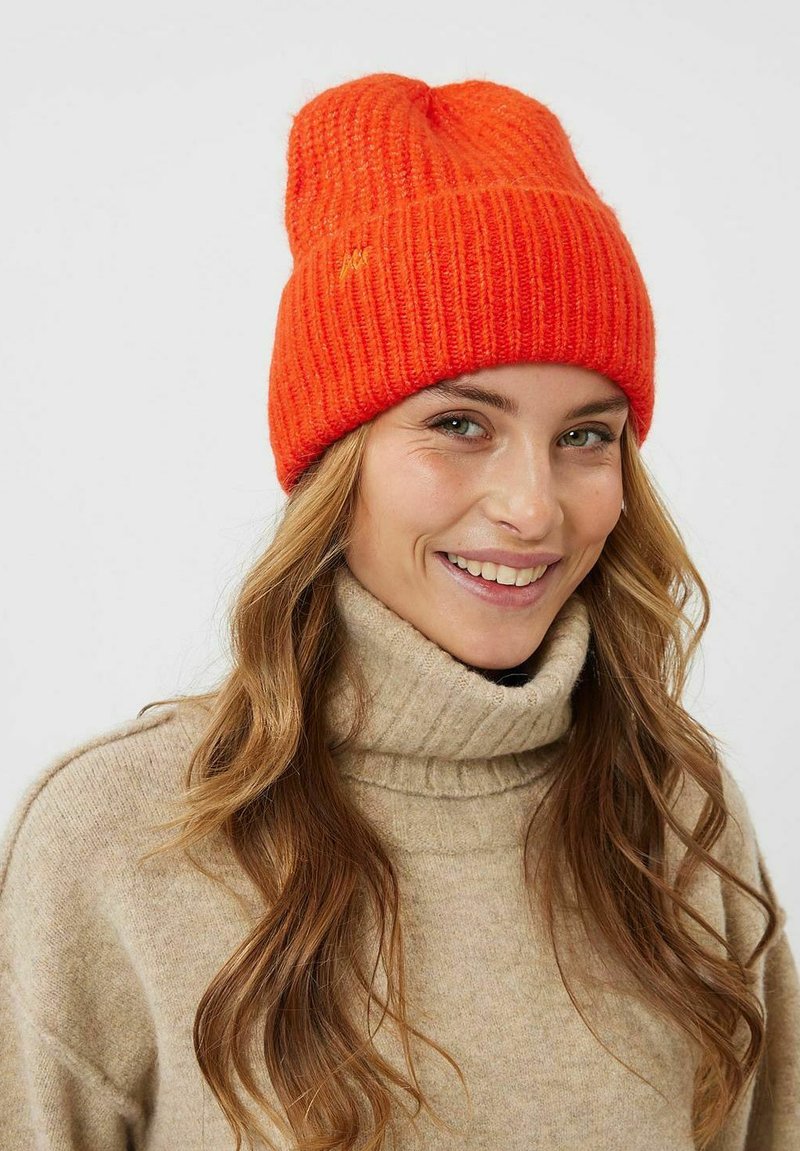 Beanie arancione lavorato a maglia con texture a coste, caratterizzato da un orlo piegato. Indossato con un maglione beige a collo alto, che mette in mostra un look casual.